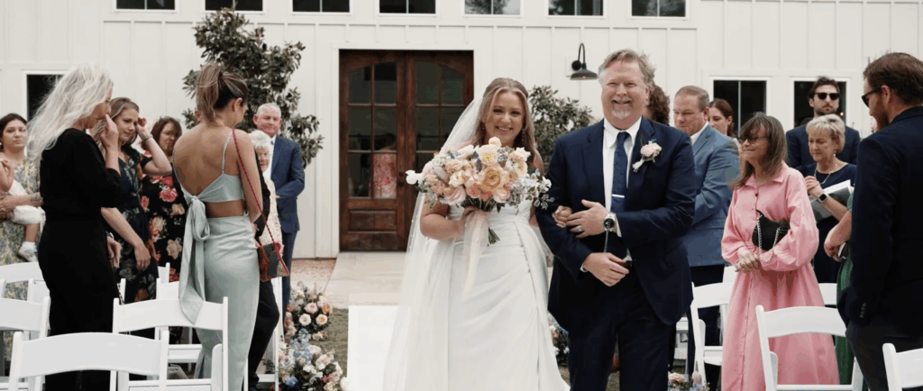 Bride walking down the aisle with her father at a wedding ceremony, surrounded by seated guests and floral decorations.