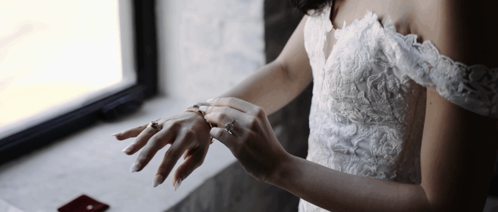 Close-up of a bride in an elegant wedding dress gently touching her bracelet, the frame highlighting the gown’s fabric and delicate details without showing her face.
