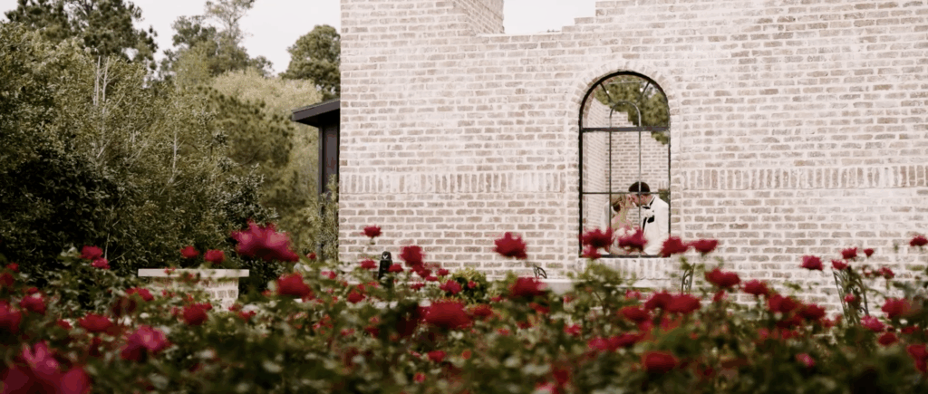 Newly married couple kissing at an outdoor wedding venue, seen through a window with soft reflections and greenery in the background.