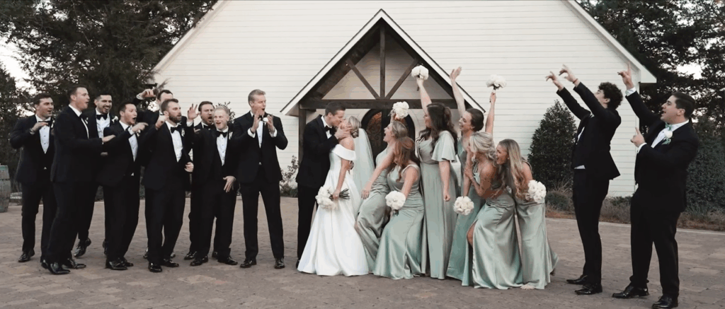 Newly married couple kisses with the bridal party on either side of them, cheering them on, at their outdoor venue.