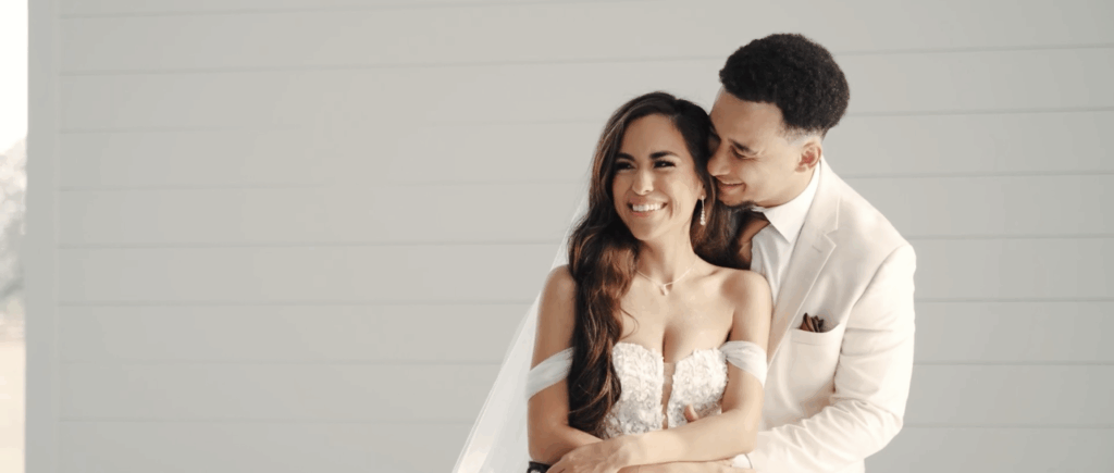 Groom wraps his arms around the bride from behind, looking at her with affection as she smiles.