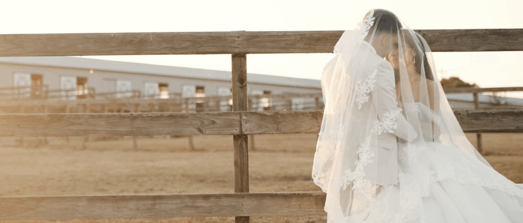 Bride and groom standing close together beneath the bride’s flowing veil in front of a rustic horse fence, creating a soft, romantic scene.