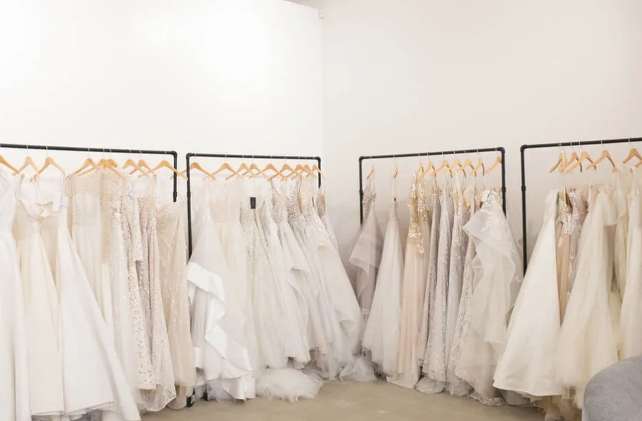 Selection of designer wedding gowns hanging on wooden racks inside Anna Bé bridal shop in Dallas, with soft natural light highlighting the ivory and lace fabrics.
