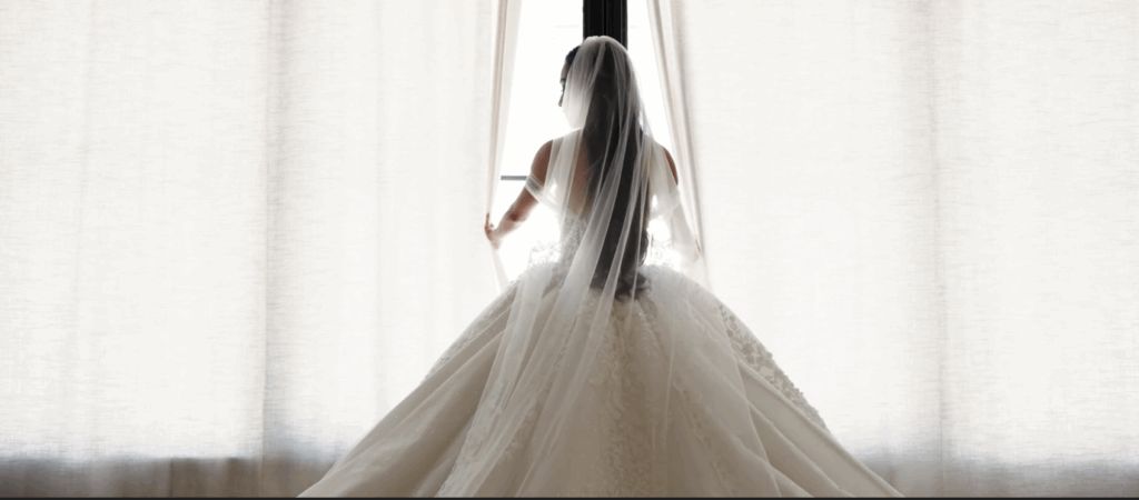 Bride in a white wedding gown standing with her back to the camera, facing a bright window, her veil and dress flowing gracefully behind her.