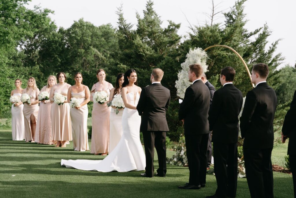 Bride and groom exchanging vows outdoors at Cinnamon Barn, with guests, bridesmaids, and groomsmen watching their ceremony.