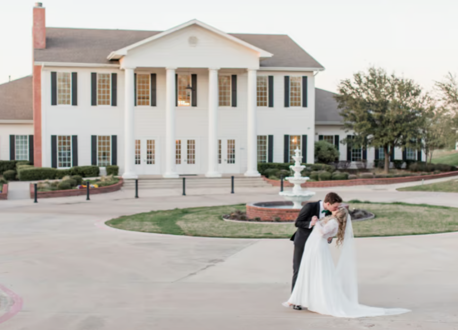 Bride and groom sharing a kiss in front of Milestone Mansion wedding venue, with the grand estate and landscaped grounds in the background.
