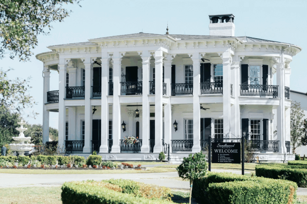 Sandlewood Manor near Houston, Texas is a grand white Southern-style mansion with tall columns, manicured lawns, and a serene pond in front.