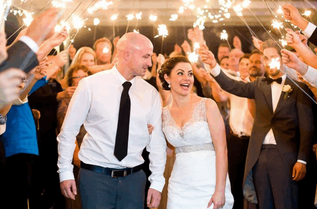 Groom looking lovingly at his bride as the two walk hand in hand through a sparkler exit, guests holding sparklers on both sides.