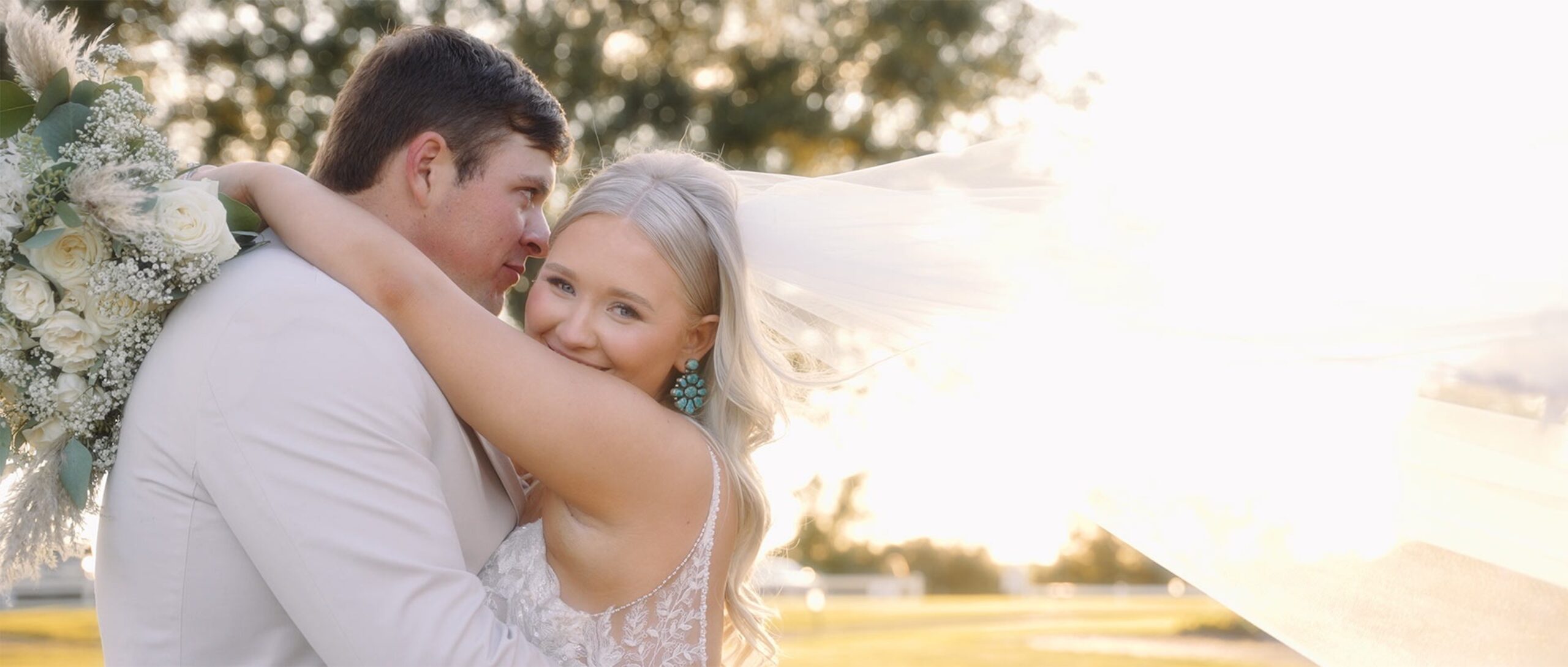 A bride smiles warmly at the camera while her groom kisses her cheek as the setting sun casts a golden glow over them.