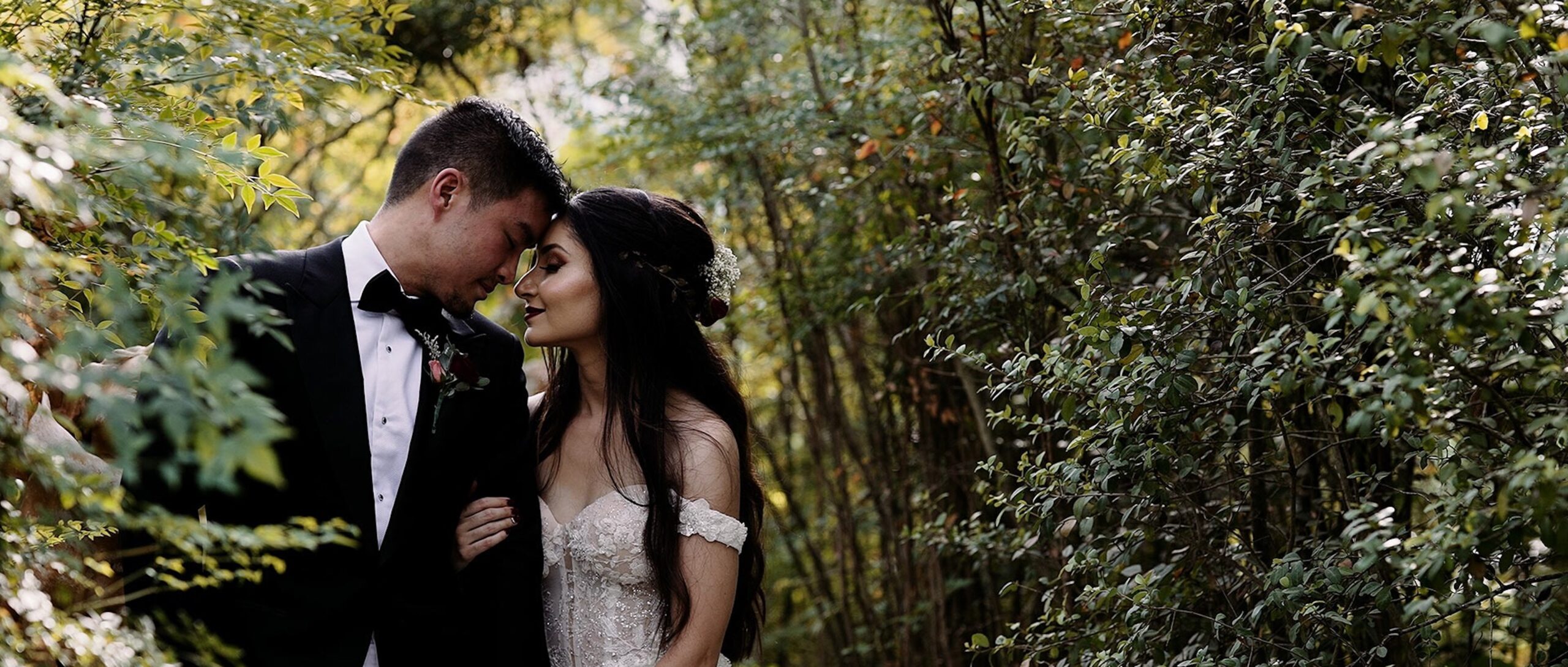Bride and groom standing close together with foreheads touching, surrounded by soft greenery, sharing an intimate outdoor moment.
