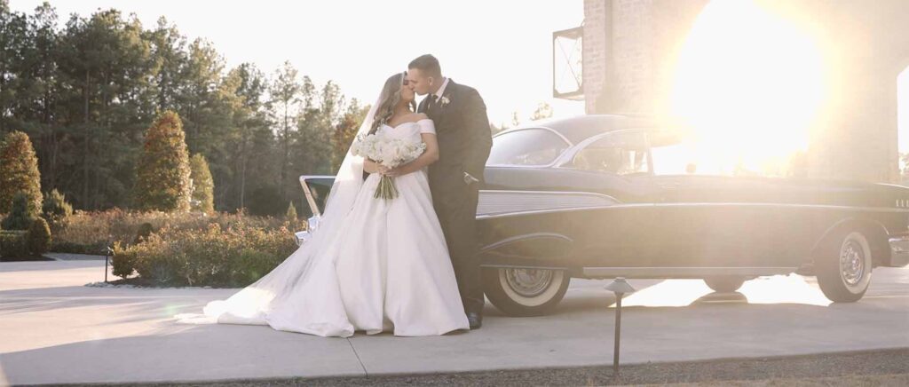 Bride and groom share a romantic kiss in front of a classic vintage car parked outdoors, capturing a timeless wedding moment.
