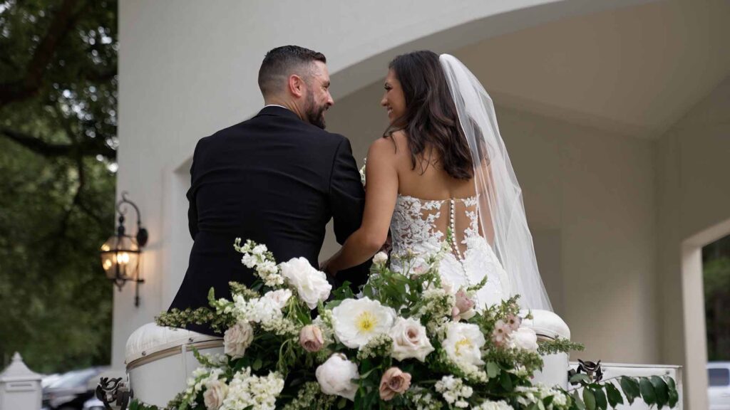 Bride and groom sit on stools outside at their venue, facing away from the camera. They are looking at each other and smiling, while we see the back detail of the bride's wedding dress.