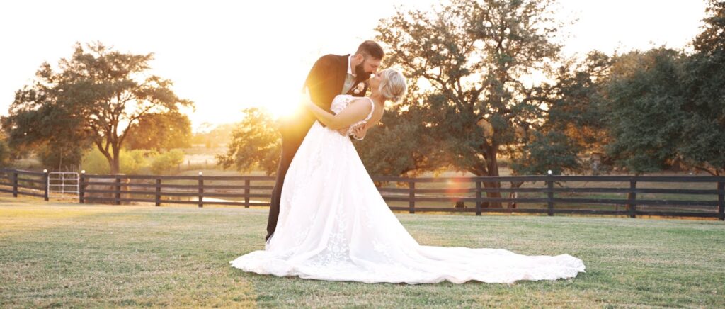 Groom dipping his bride for a kiss in front of a rustic fence at an outdoor Houston wedding venue, with greenery in the background.
